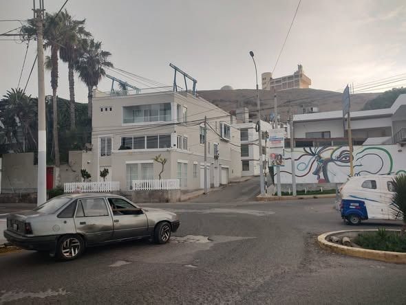 Escena callejera con edificios, palmeras y un coche girando. Al fondo se ve una colina.