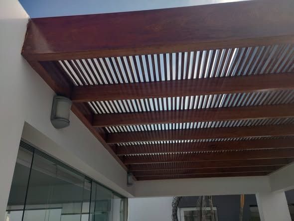 Pérgola de madera con lamas sobre patio, adosada a edificio blanco.