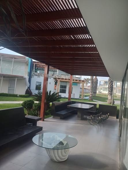 Patio exterior con pérgola de madera, asientos negros y mesa de cristal.