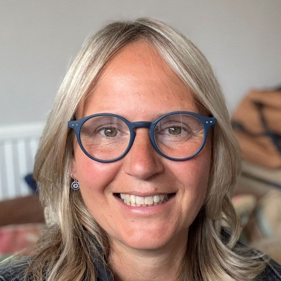 Woman with blonde hair, round blue glasses, smiling, inside.