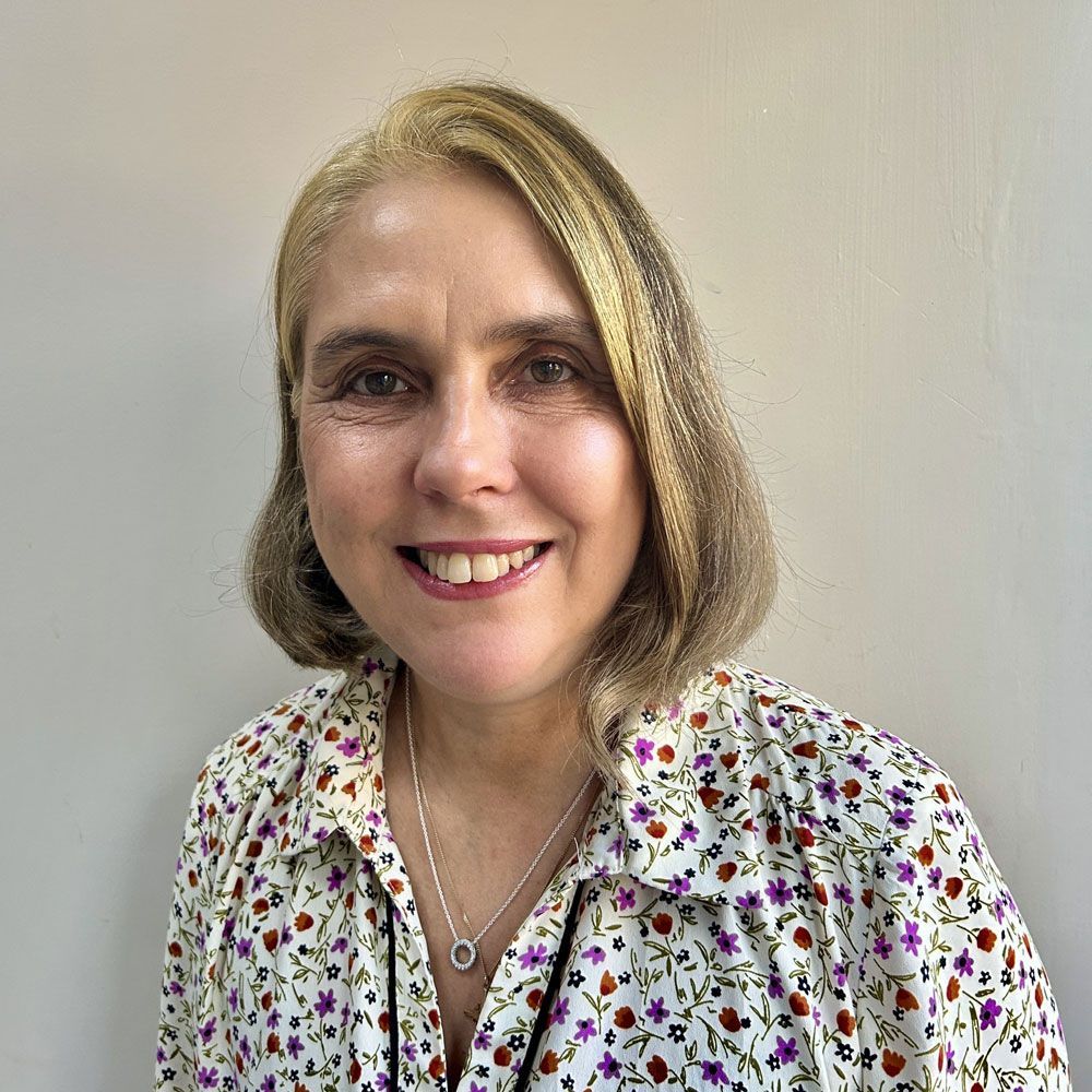 Woman with light brown hair, smiling, wearing floral shirt, silver necklace, indoors.