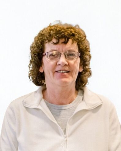 Woman with curly brown hair, glasses, smiling, wearing a white jacket over a gray shirt.