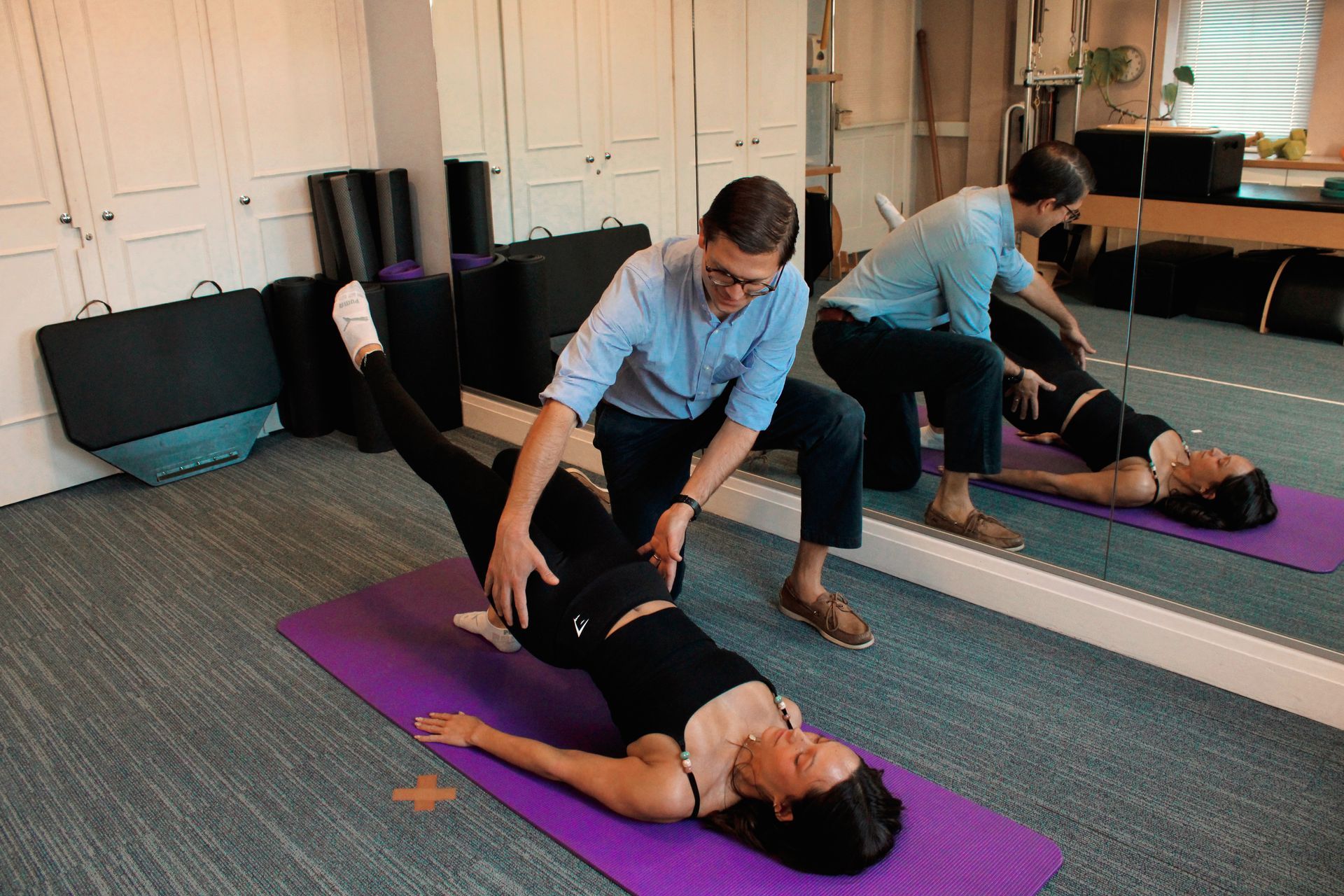 Two people doing exercise with assistance on a purple mat in a studio with a mirror.