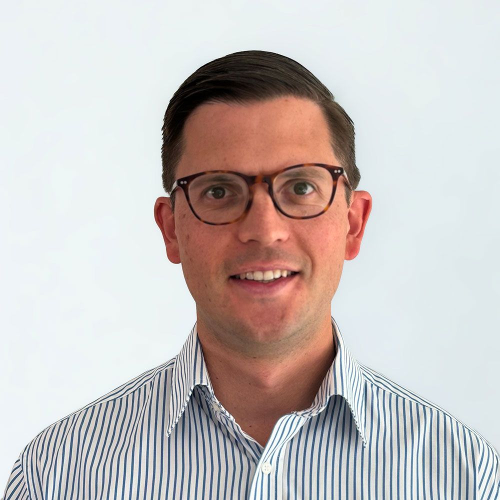 Man with glasses, wearing a striped shirt, smiling in front of a white background.