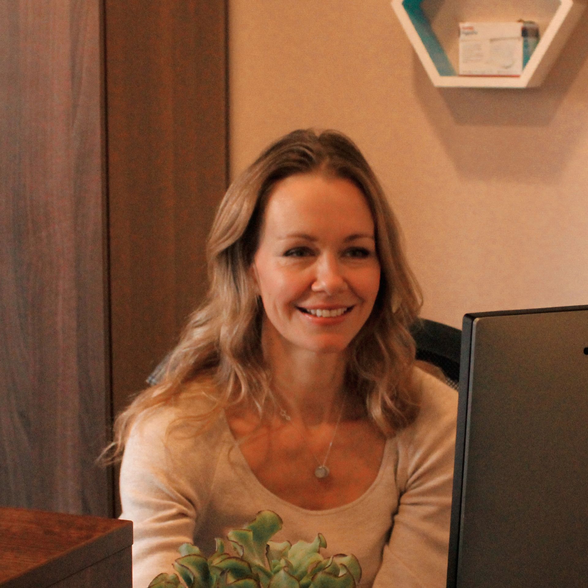 Woman with long hair smiles, working at a computer in an office setting.