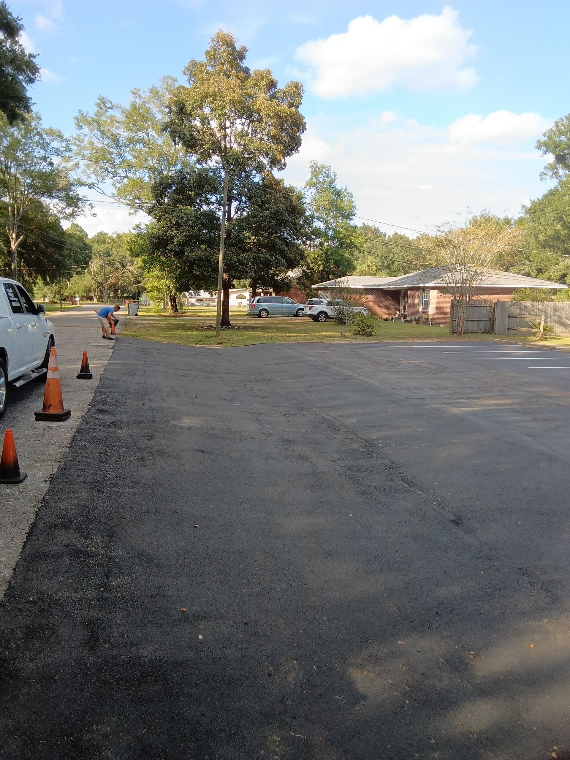 Newly paved asphalt, orange cones, and a person in the distance near trees and homes under a blue sky.