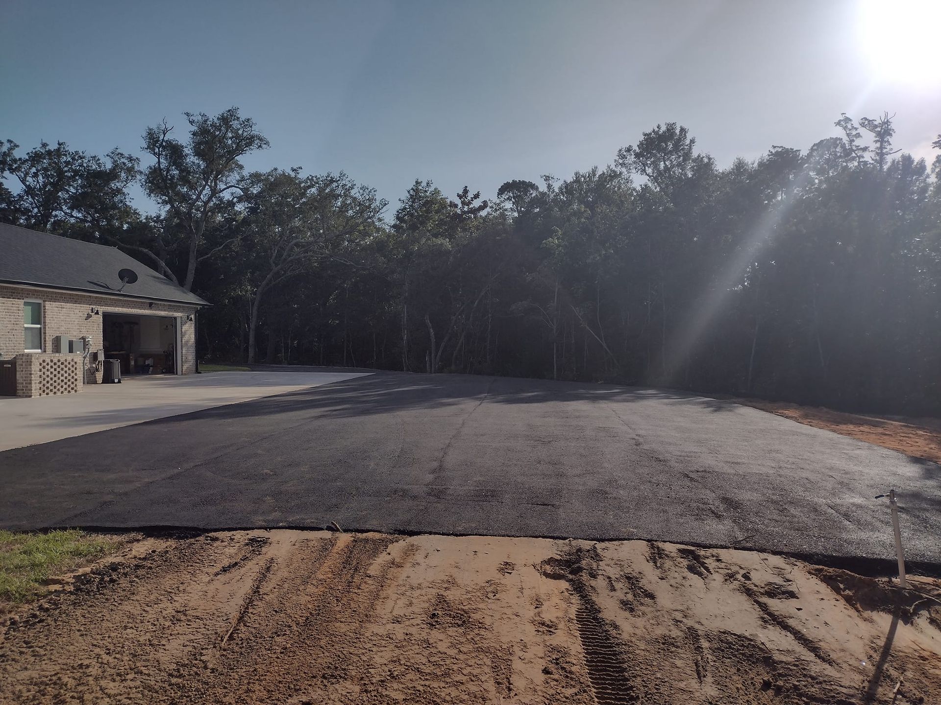 Newly paved driveway, dark asphalt, in front of a house, set against a backdrop of trees under a blue sky.
