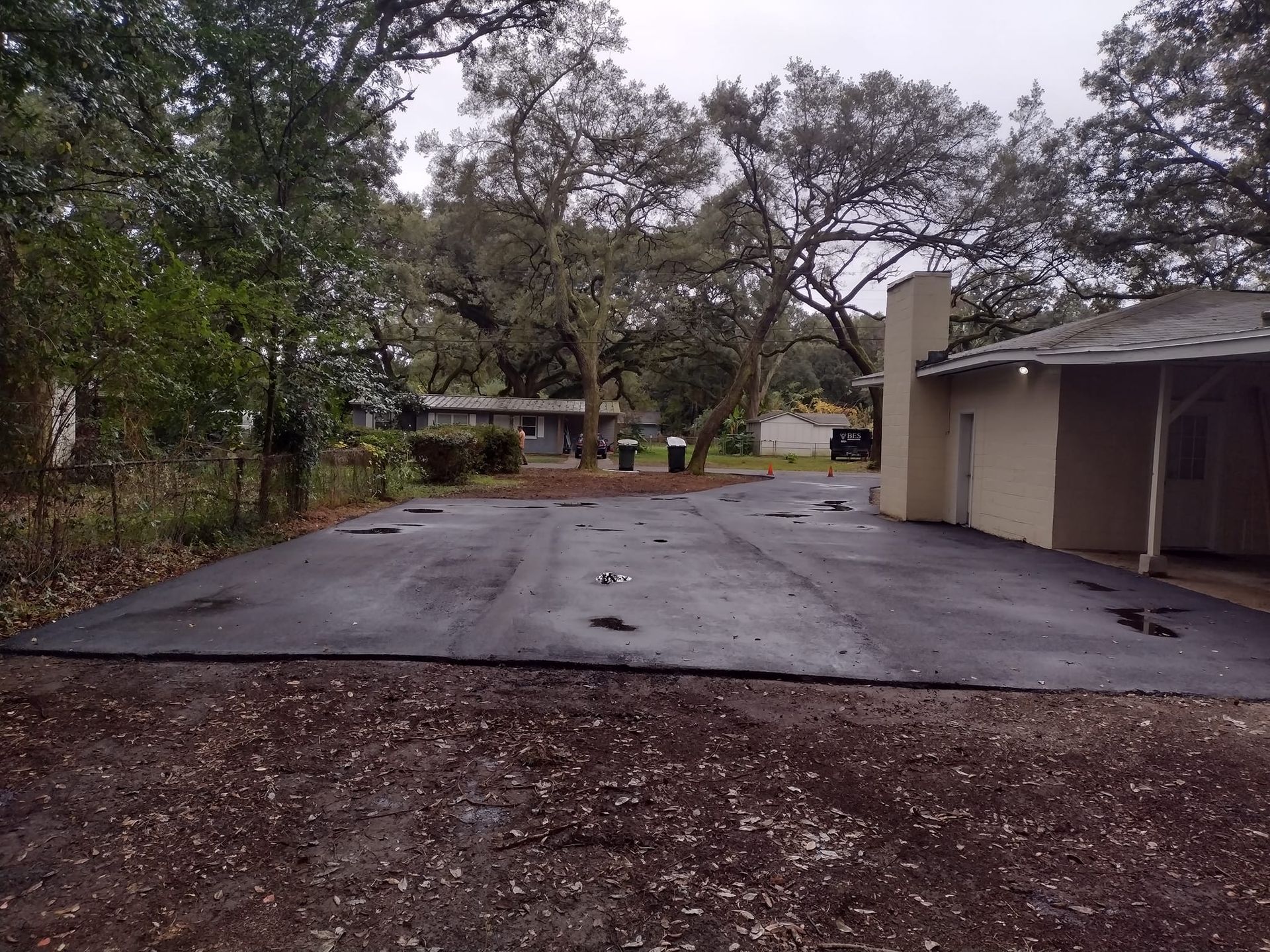Black asphalt driveway with a beige building, surrounded by trees.
