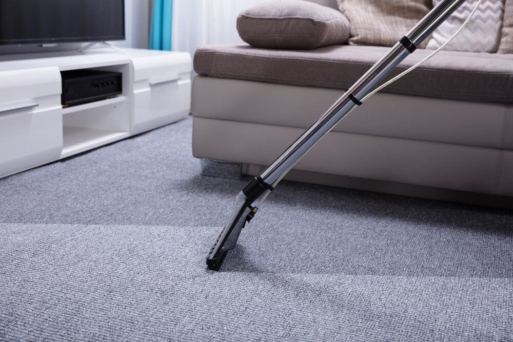 Vacuum Cleaner Nozzle Cleaning Gray Carpet Beside a Beige Sofa — Broadbeach Carpet Cleaning In Broadbeach, QLD