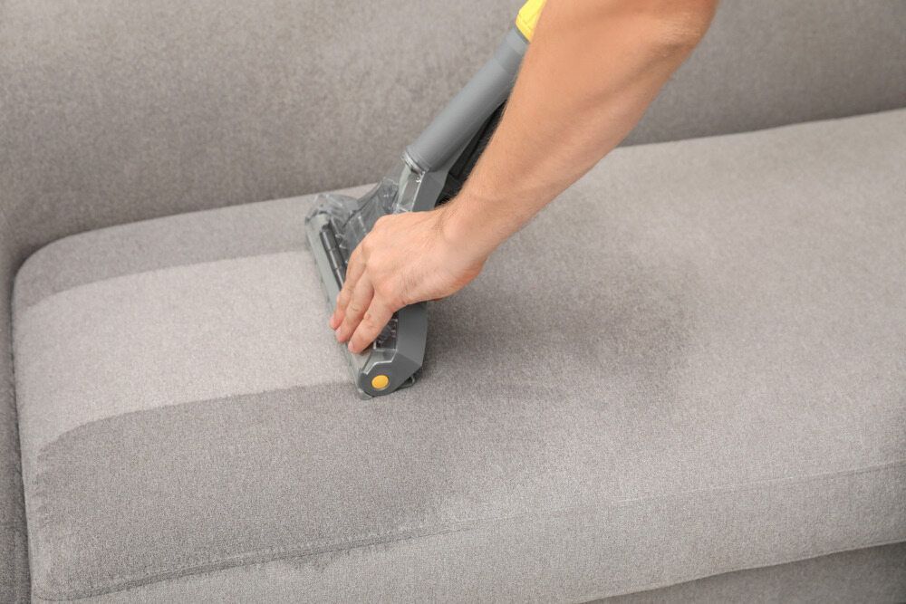 Hand Using a Vacuum Cleaner on A Gray Sofa Cushion — Broadbeach Carpet Cleaning In Broadbeach, QLD