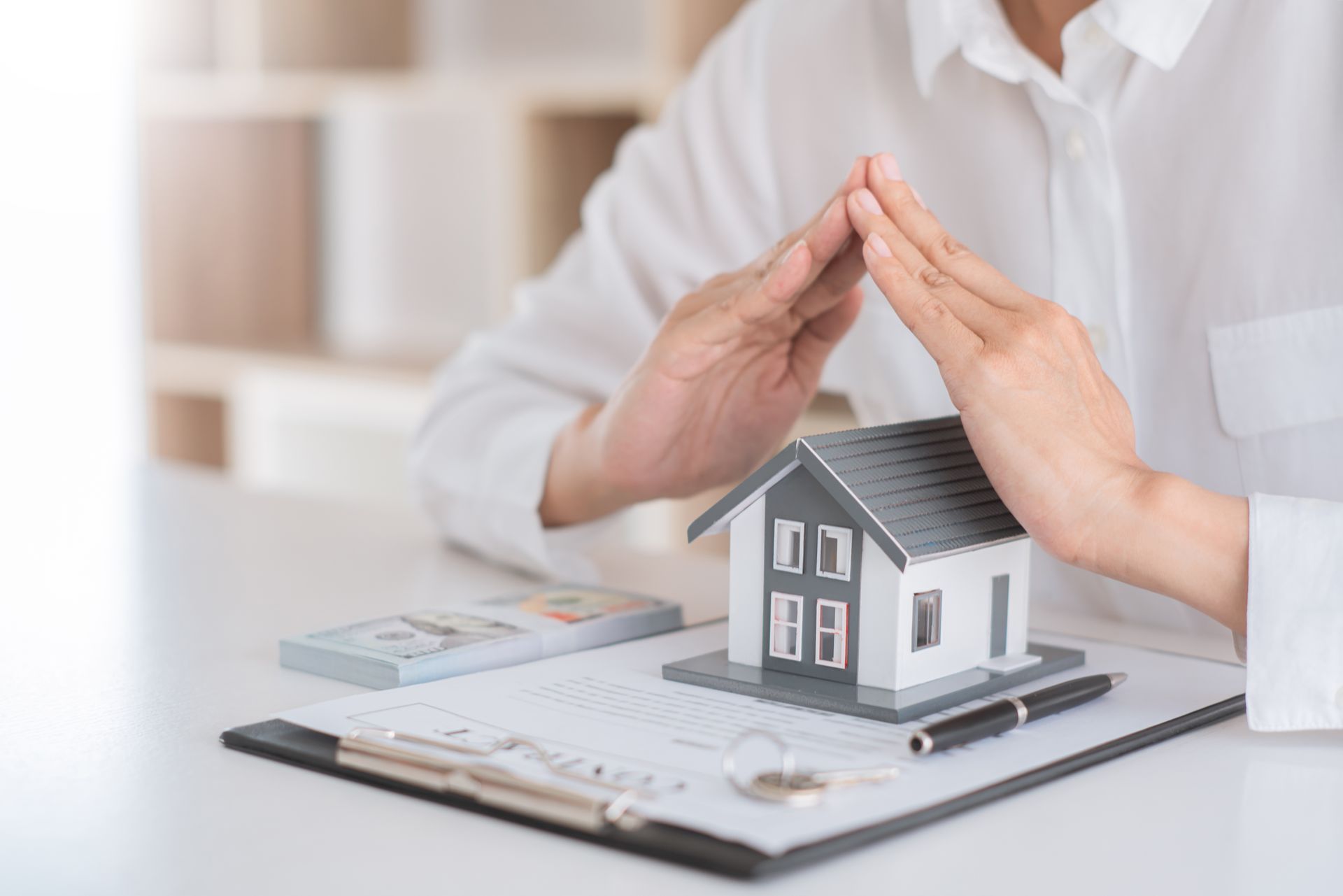 Hands protecting a miniature house on a desk with documents, money, and a pen.