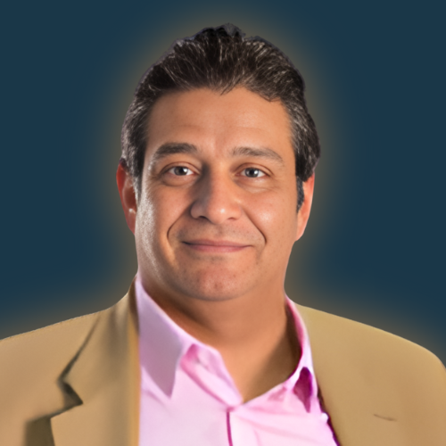 A man wearing a tan jacket and a pink shirt smiles for the camera