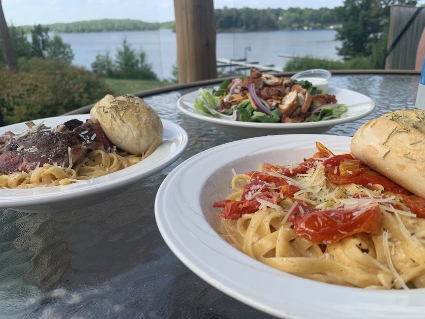 Three plates of pasta and bread on an outdoor table overlooking a lake