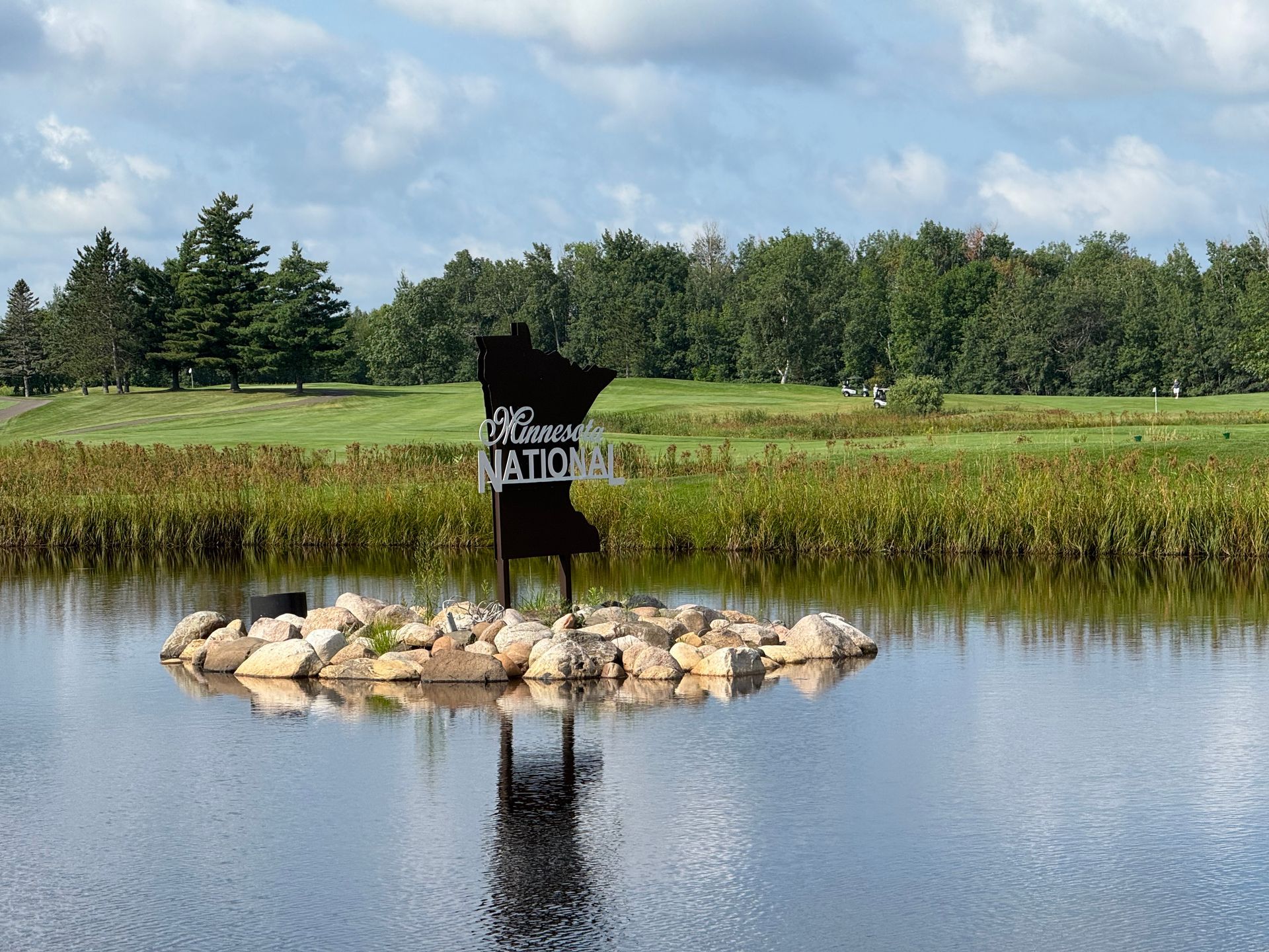 Small pond with a rock island and black “Welcome to Natchitoches” sign, framed by trees and grass.