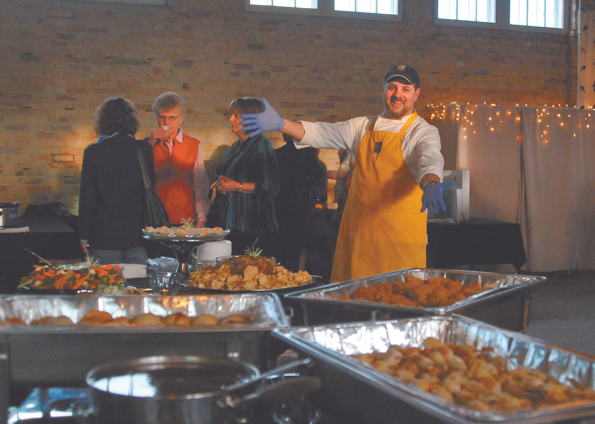 Buffet table with trays of food as people serve guests in a brick-walled hall