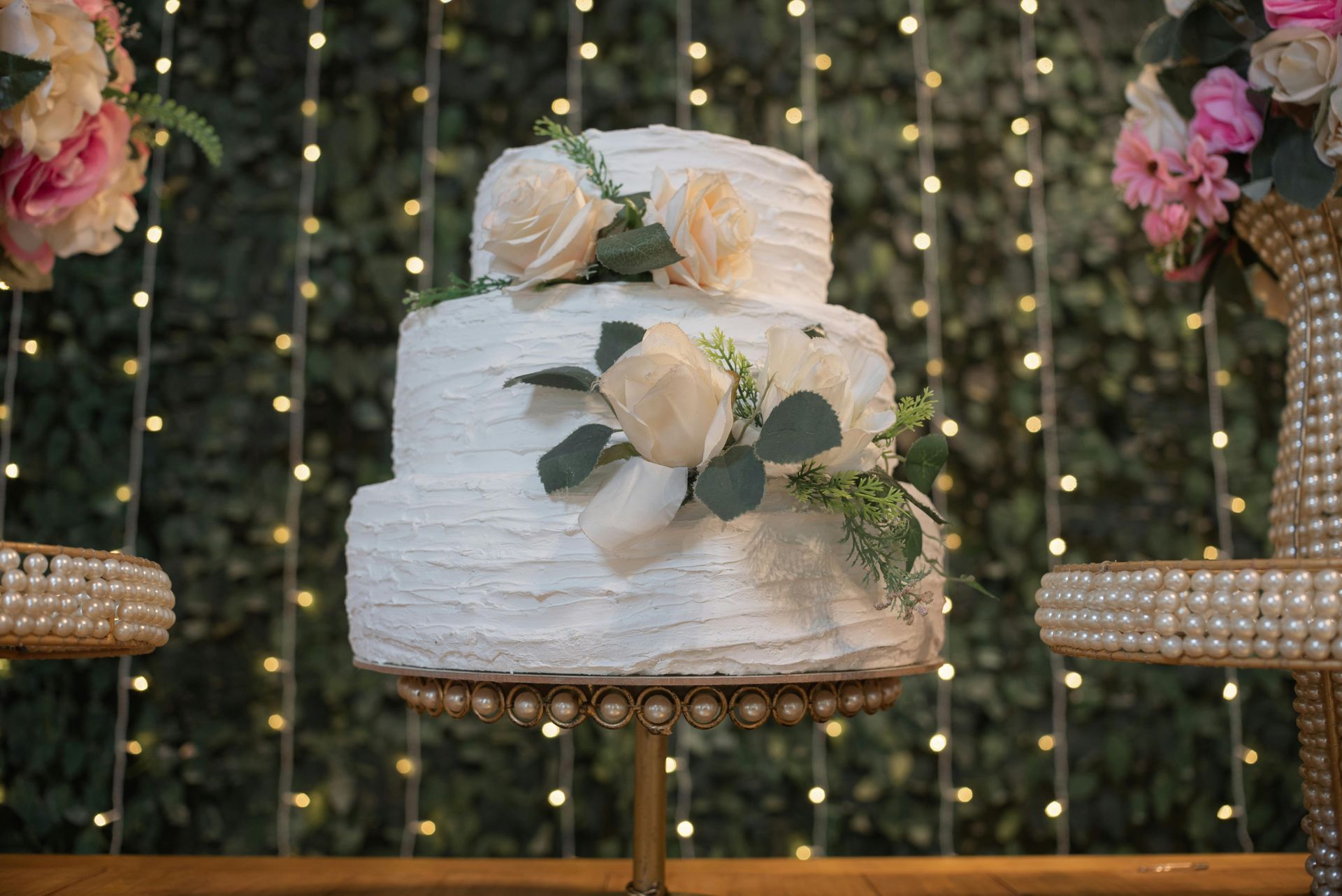 White two-tier wedding cake with cream roses and greenery, displayed on a decorated stand with fairy lights behind it