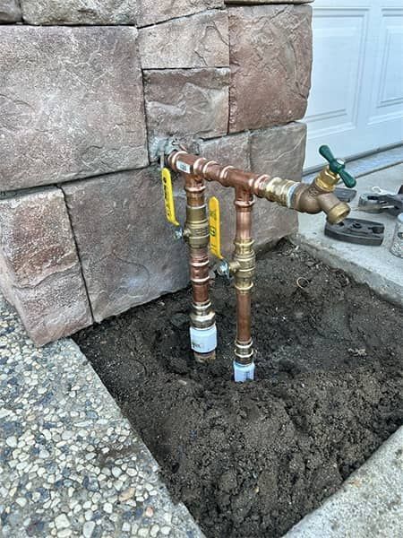 Exposed copper pipes and shutoff valves in a dirt trench beside a stone wall and garage door