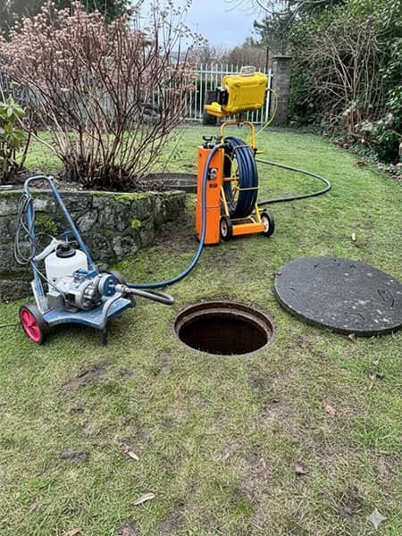 Yellow drainage machine and hoses on a lawn beside an open manhole cover.