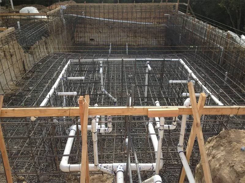 Construction site with rebar foundation grid and white PVC plumbing pipes in a dug-out foundation pit