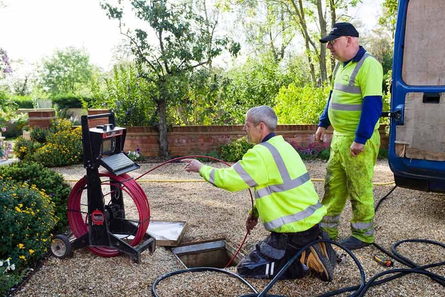 Two workers in yellow hi-vis gear inspect a red pipe-cleaning machine in a garden area.