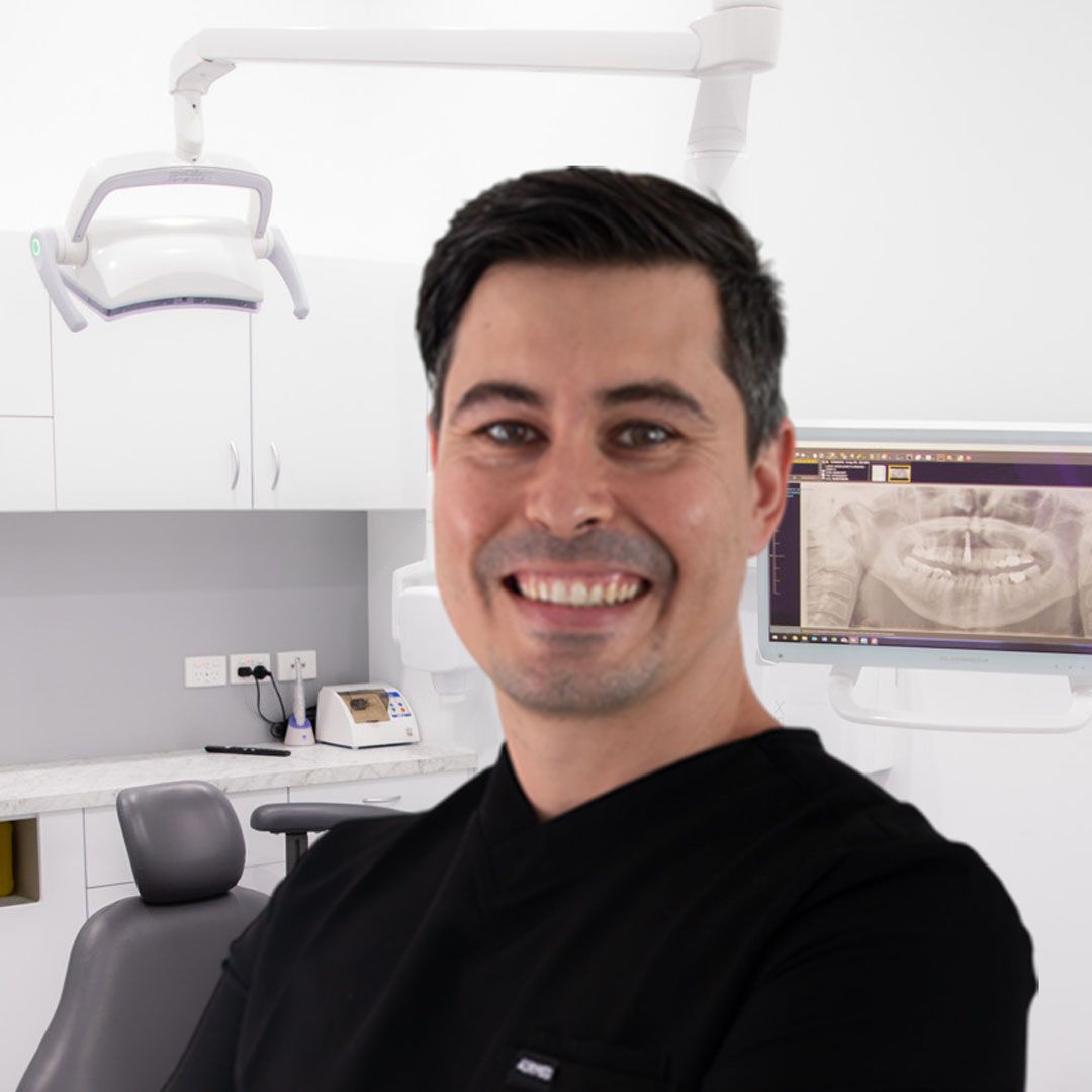 Smiling man in black top at a dental office. Dental chair and X-ray visible.