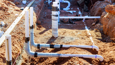 White PVC pipes and fittings installed in a dirt trench at a construction site.
