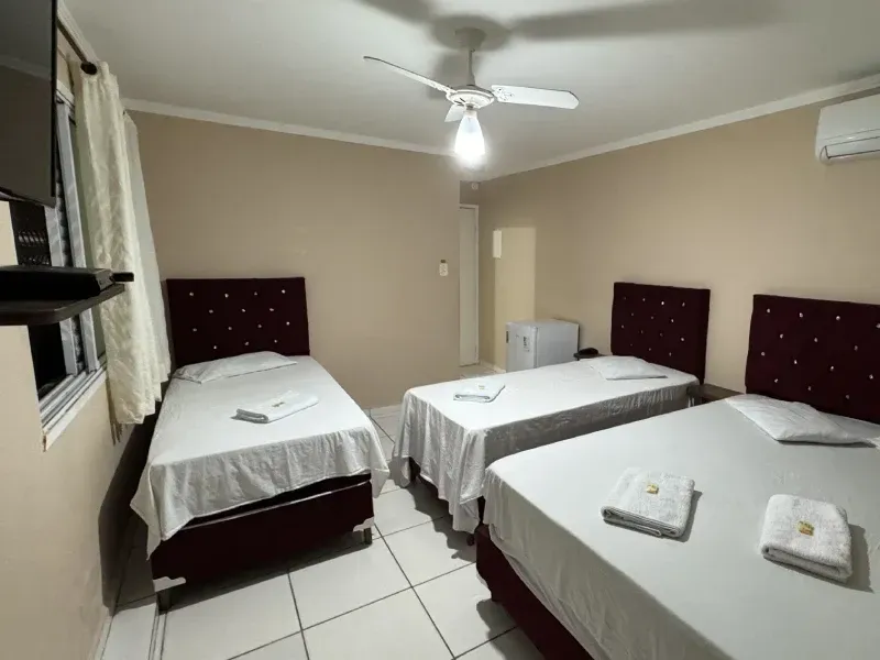 Um quarto de hotel com três camas, cabeceiras estofadas escuras e roupa de cama branca, com ventilador de teto e piso de azulejo.