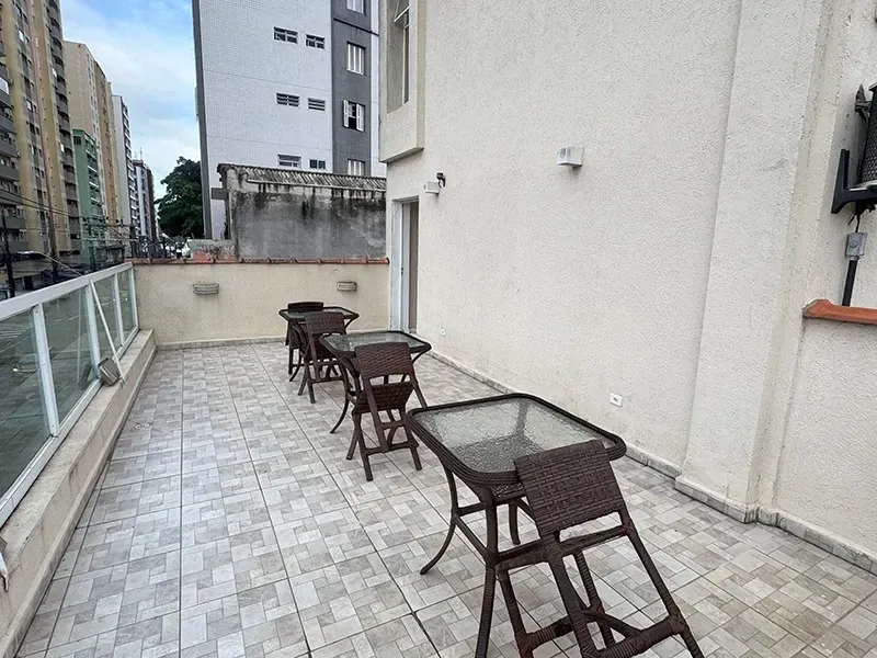 Uma área de varanda externa com três pequenas mesas quadradas e cadeiras de vime dispostas sobre um piso de azulejos claros.