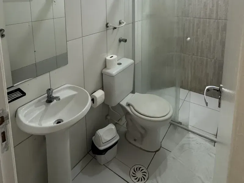 Um pequeno banheiro com azulejos brancos, equipado com pia de coluna, vaso sanitário e box de chuveiro envidraçado.