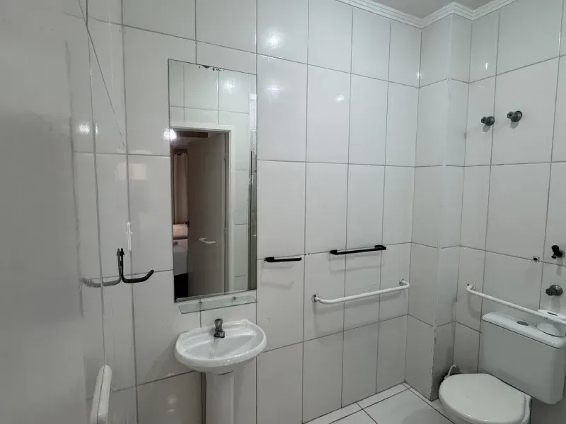 Um banheiro acessível, amplo e iluminado, com azulejos, equipado com lavatório com pedestal e espelho, e vaso sanitário com barras de apoio.