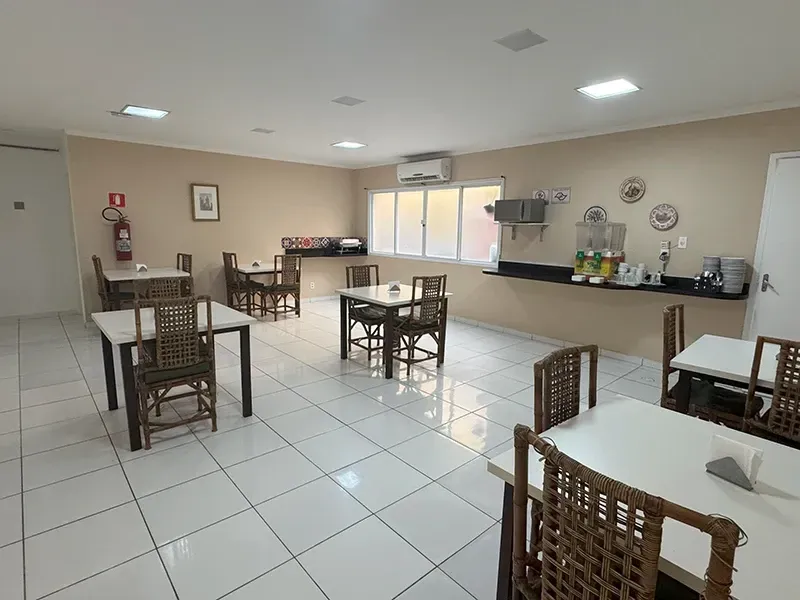 Uma sala de café da manhã de hotel bem iluminada, com fileiras de mesas quadradas e cadeiras de vime sobre um piso de azulejos brancos.