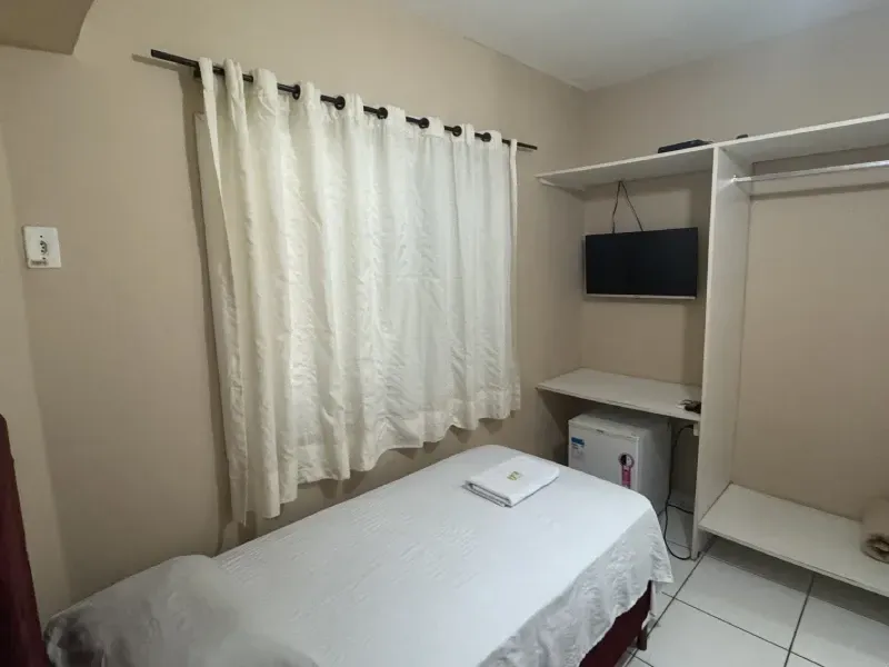 Um quarto pequeno, em tons neutros, com uma cama de solteiro, uma janela com cortinas brancas, uma TV fixada na parede e um armário aberto.