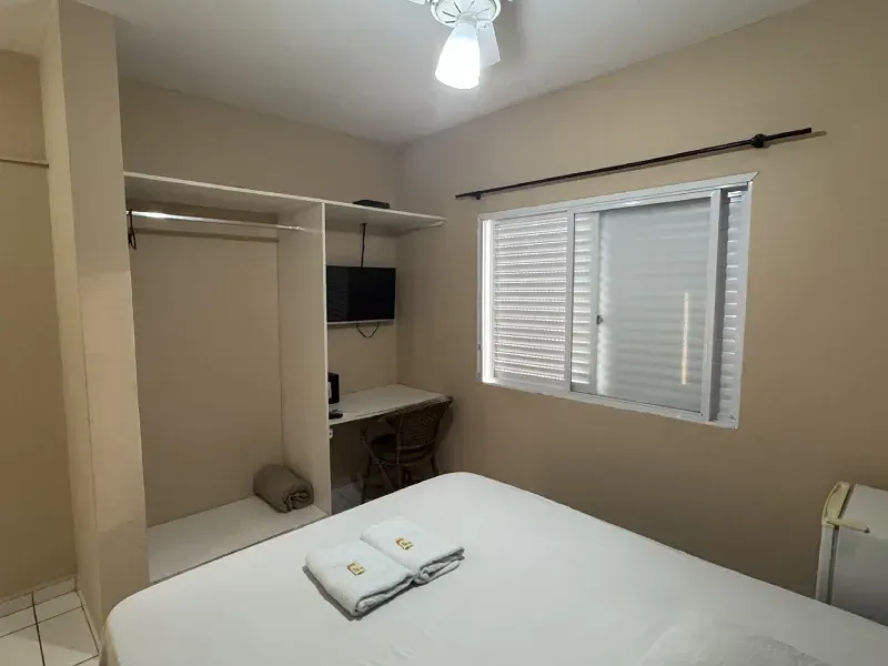 Um quarto de hotel com uma cama, um armário aberto, uma pequena escrivaninha com televisão e uma janela com persianas brancas.
