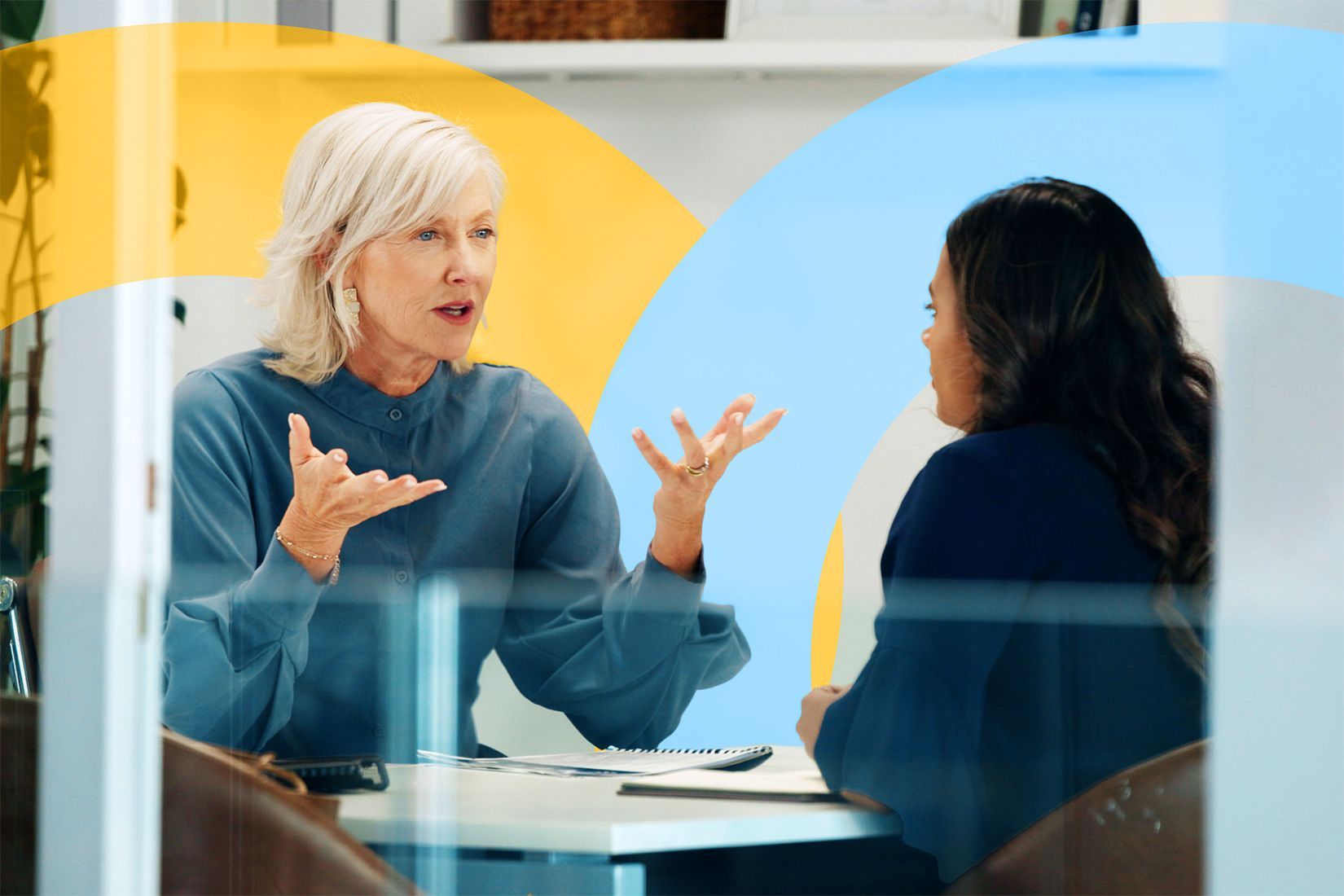One businesswoman coaching a junior staff member, her hands are raised and she is leaning forward.