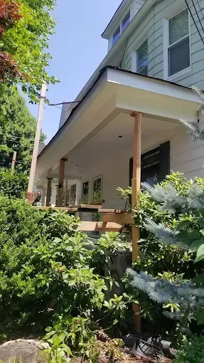 A white house with a porch surrounded by trees and bushes on a sunny day.