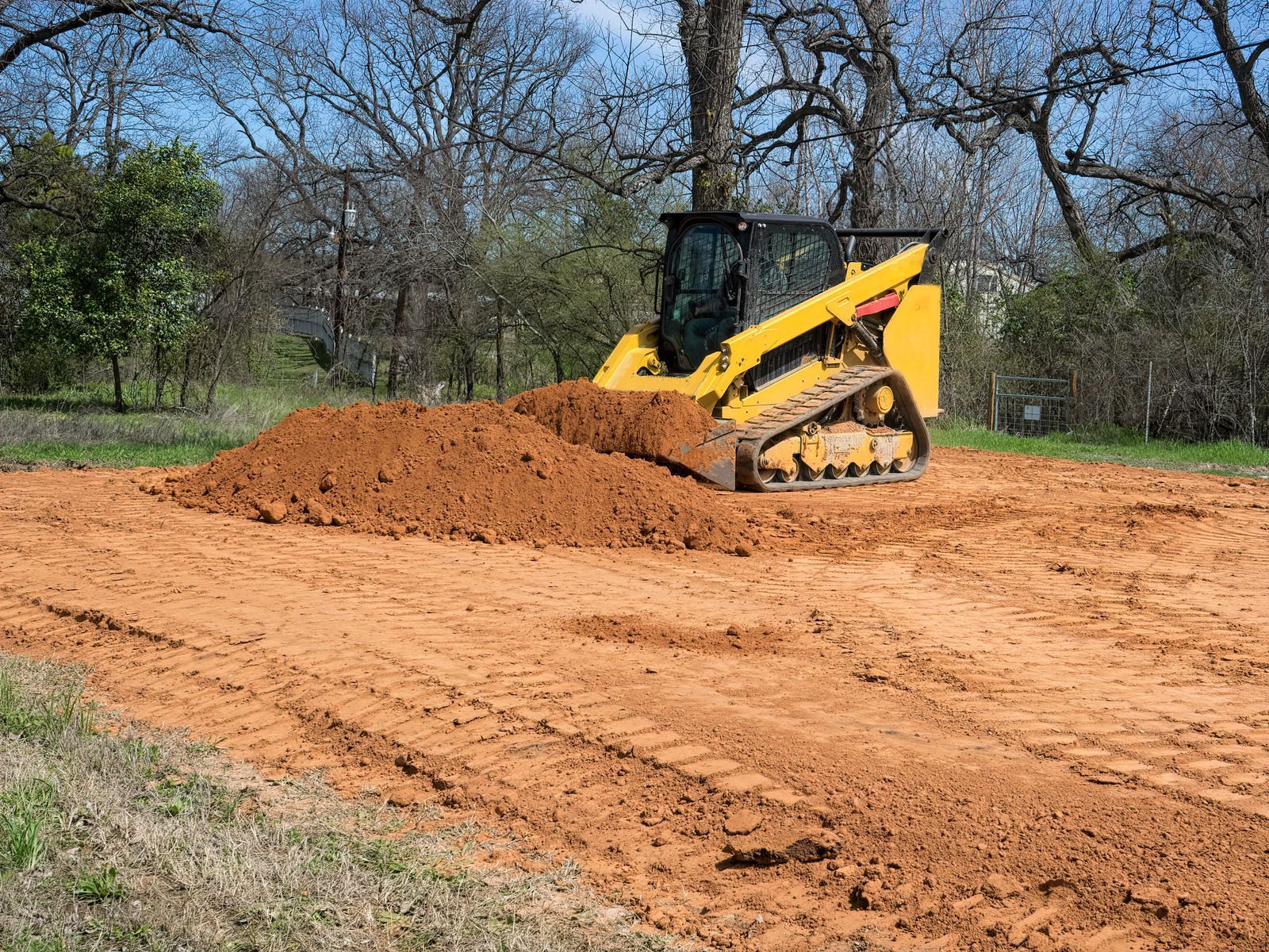 Yellow track loader moving a pile of reddish-brown dirt in an outdoor setting.