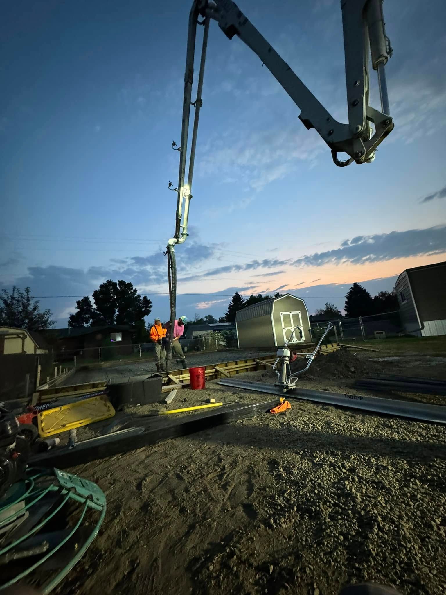 Concrete pump pouring concrete on a gravel-covered construction site with a shed and trees in the background at dusk.