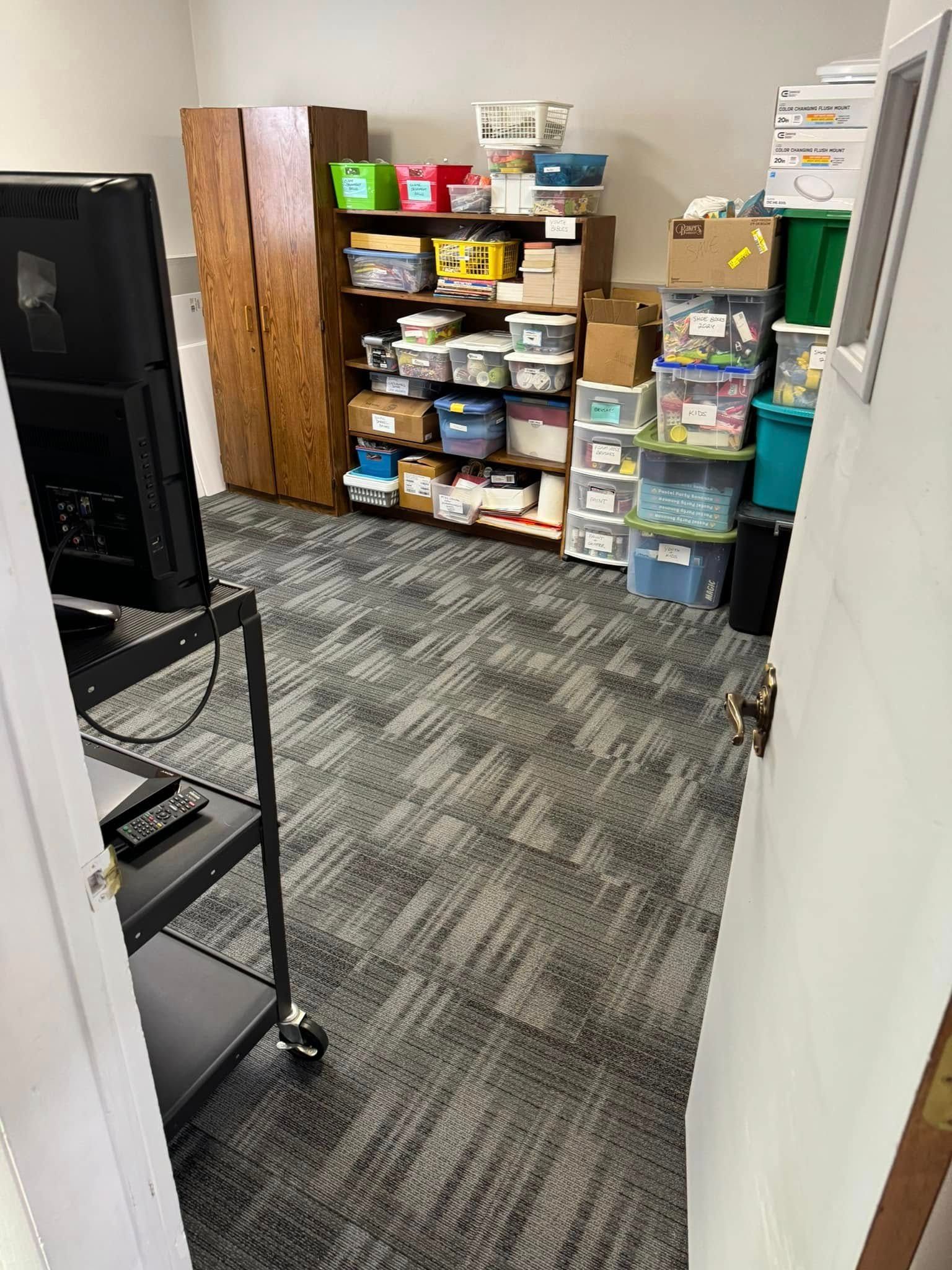 A cluttered room with shelves full of boxes, a cabinet, and a black cart. Gray carpet covers the floor.