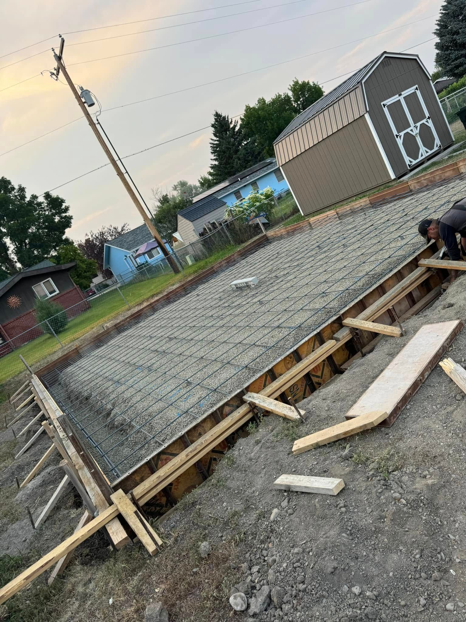 Freshly poured concrete in wooden formwork. A person works on the edge. Shed in background.