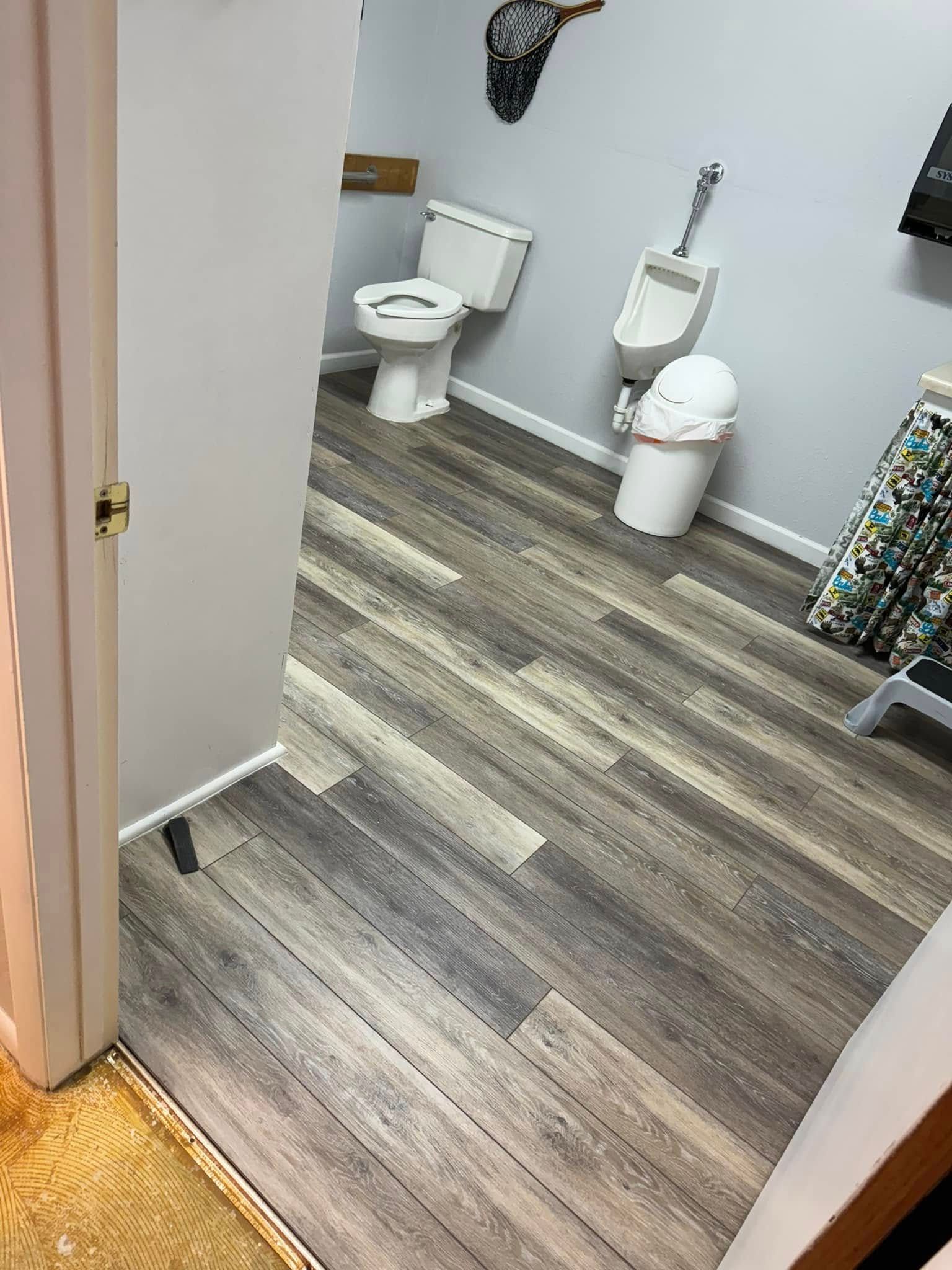 Bathroom with a toilet, urinal, and waste bin; gray wood-look flooring.