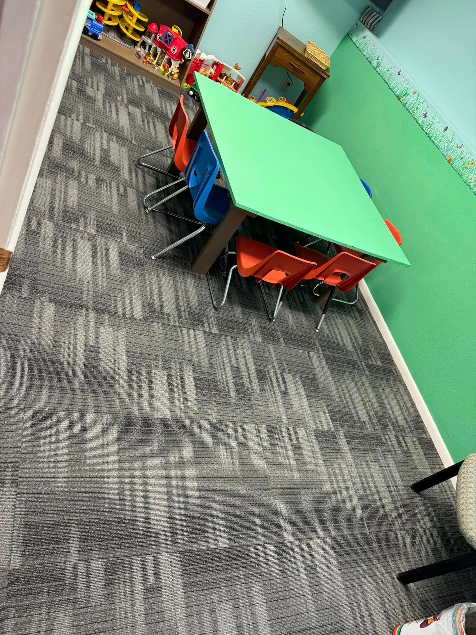 Classroom with a green square table, colorful chairs, and textured gray carpet.