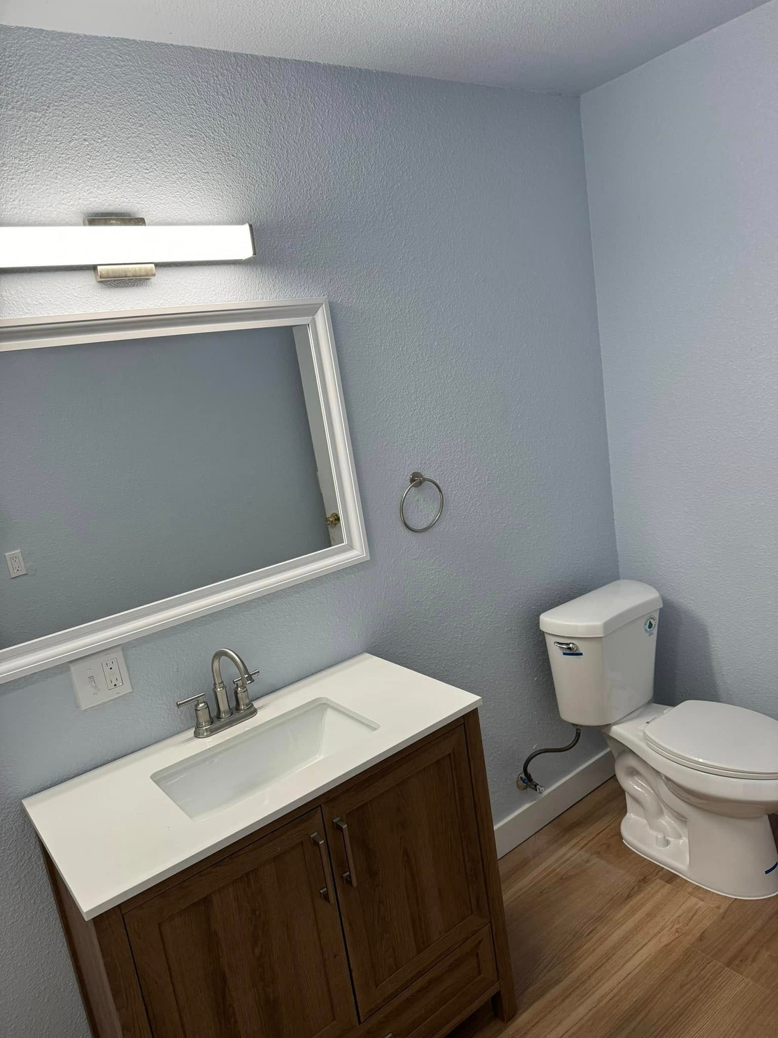 Bathroom with blue walls, wood vanity, white toilet, and mirror with light fixture.