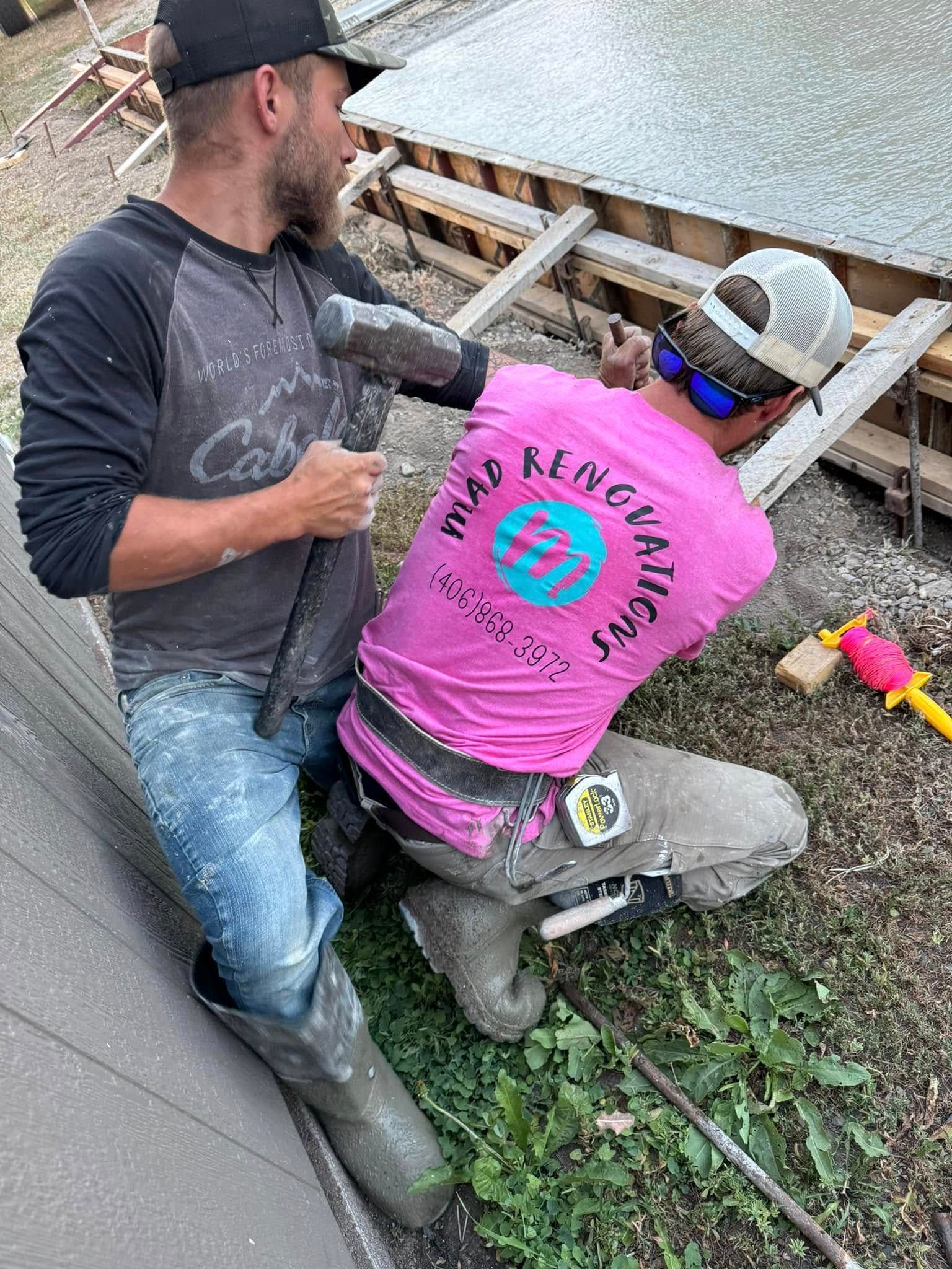 Two construction workers on a roof, one hammering wood while the other assists.