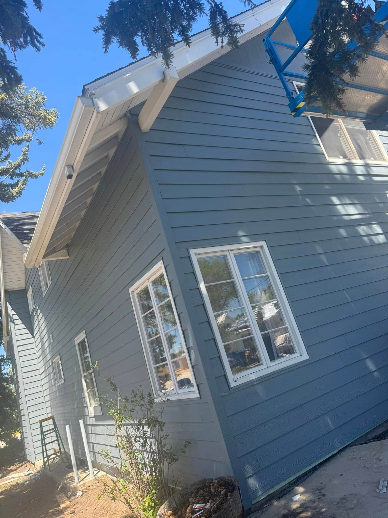 Blue siding on a house with white trim and windows under a blue sky, with some tree branches overhead.