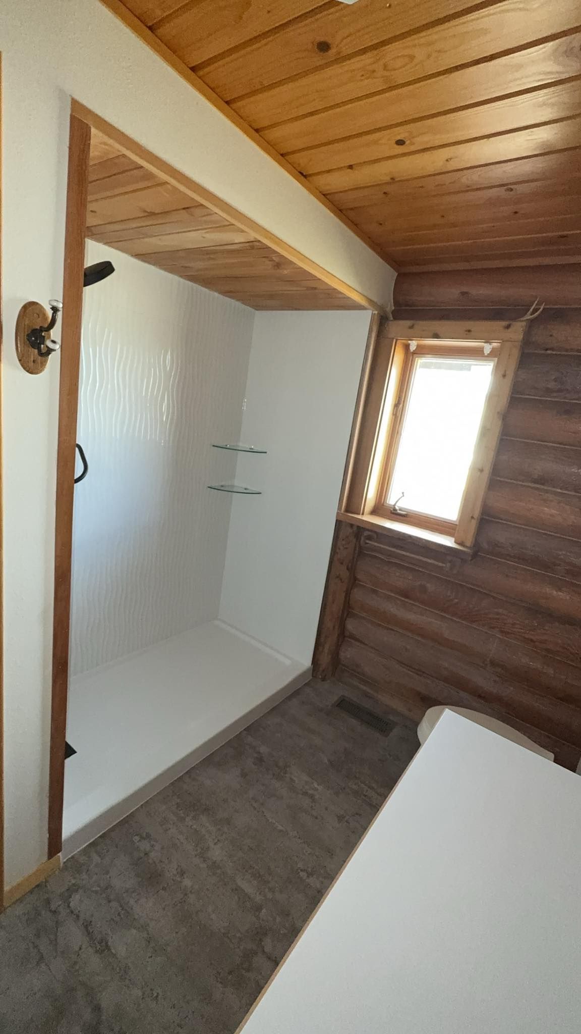 Shower in small, rustic bathroom with a window; white walls, wood trim, and a wood ceiling.