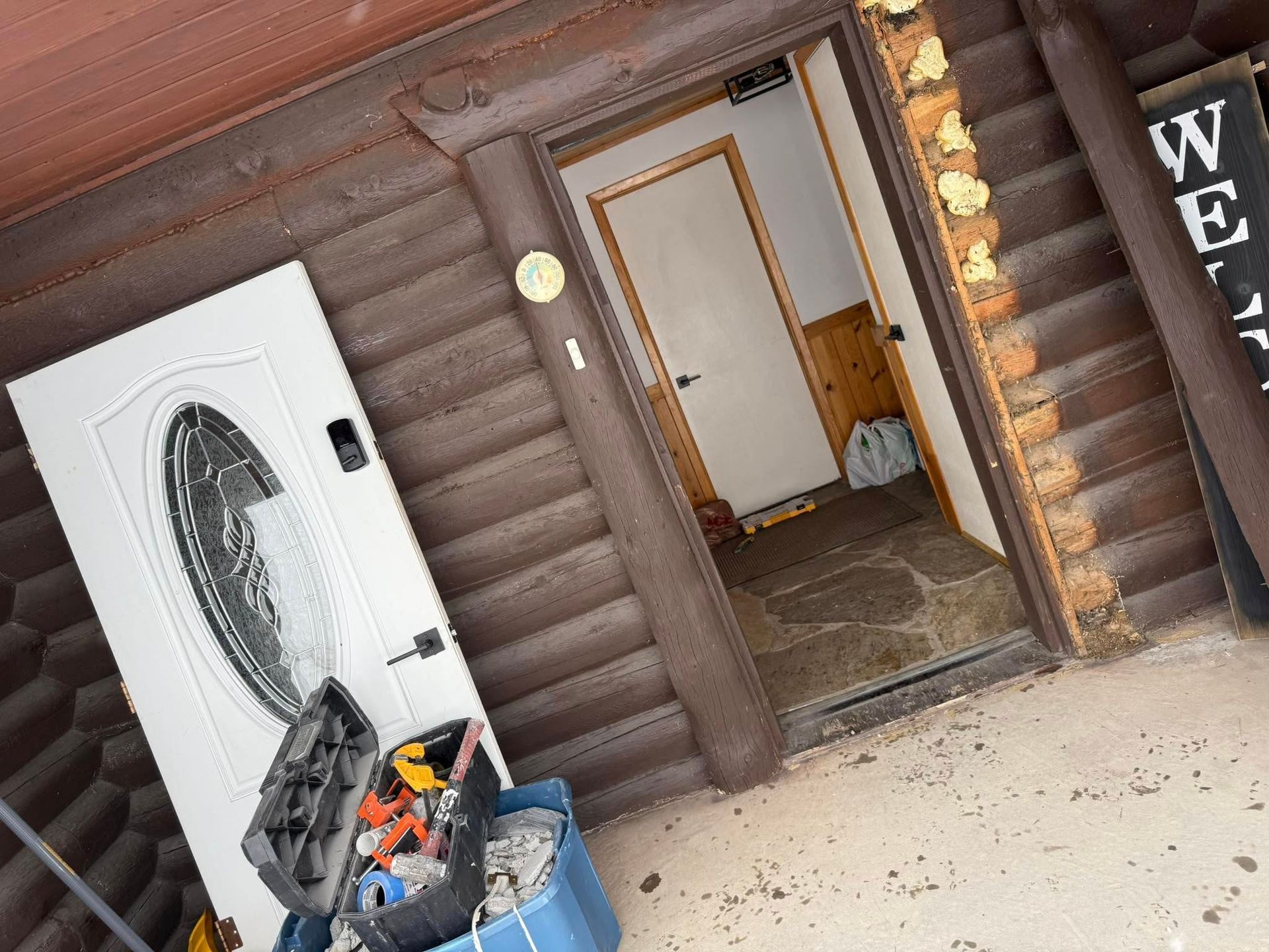 Exterior of a cabin; a new white door leans against the building. Tools and an open doorway are visible.