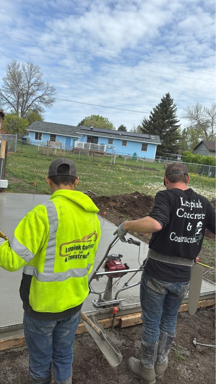 Two workers smoothing wet concrete. One in a yellow vest, the other operating a power trowel, cloudy sky in the background.