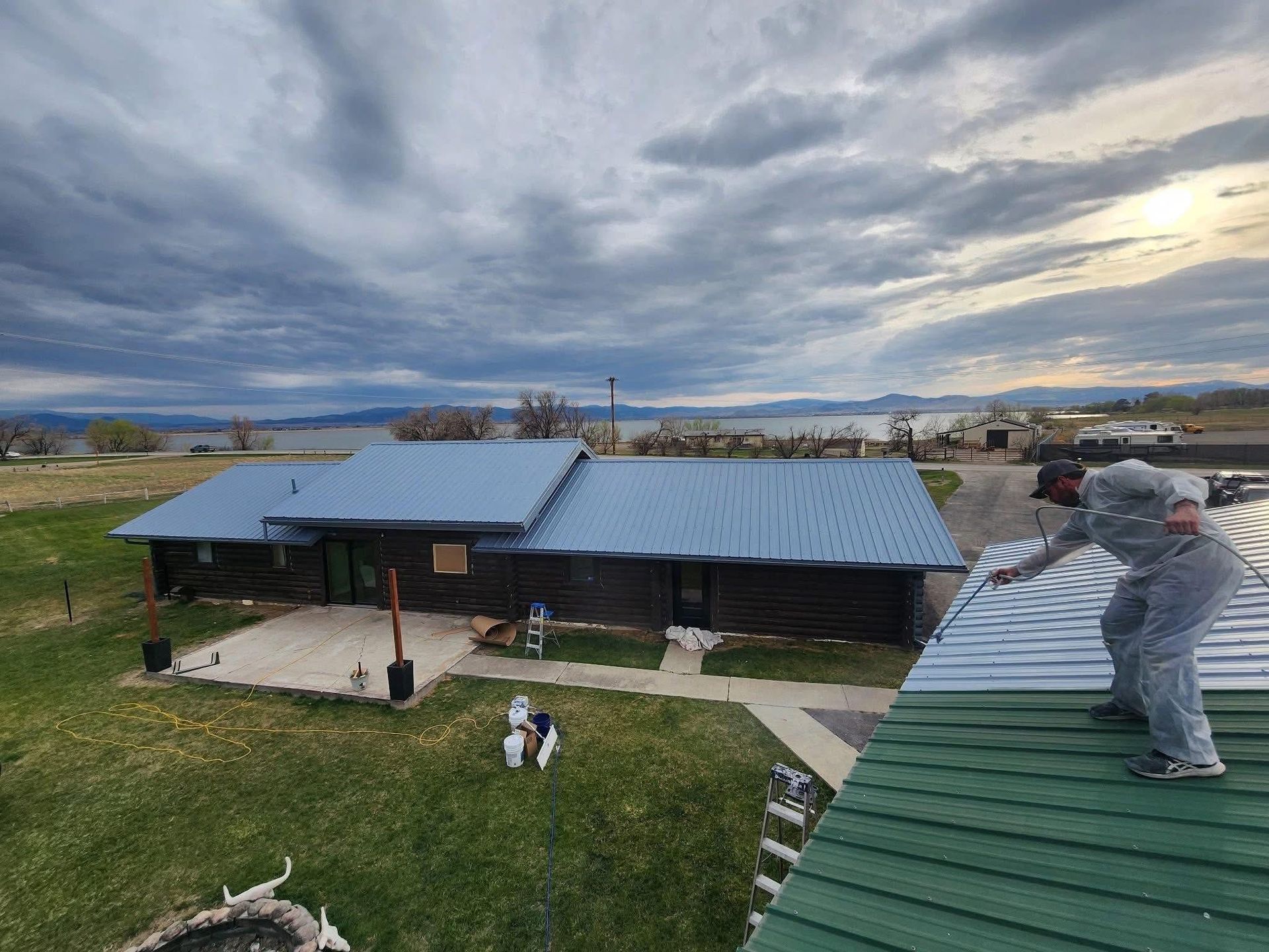 Man on a green roof pointing toward a blue metal roof on a dark wooden house near a lake under a cloudy sky.