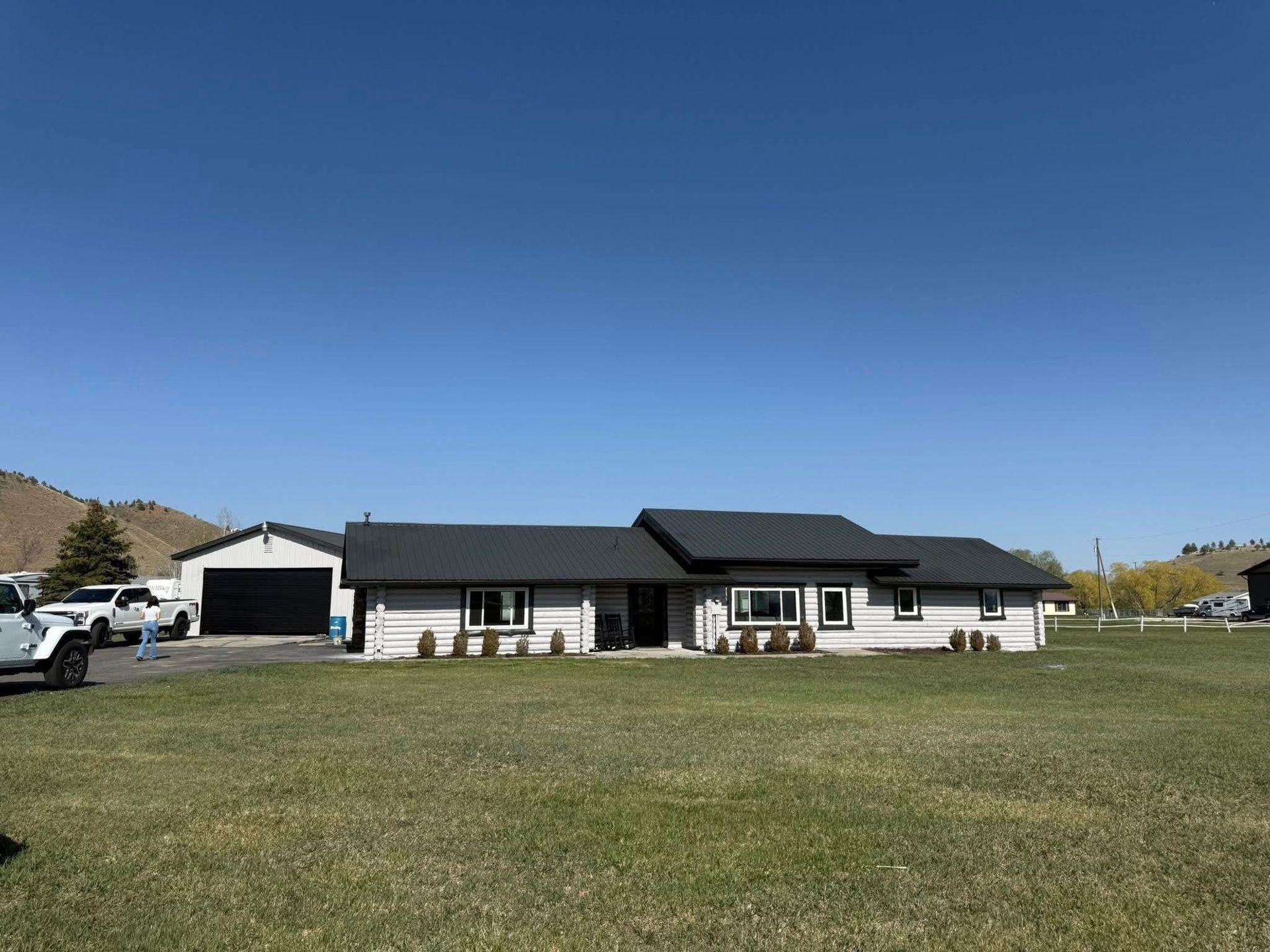 A one-story house with a black roof and white siding, sitting on a green lawn on a sunny day.