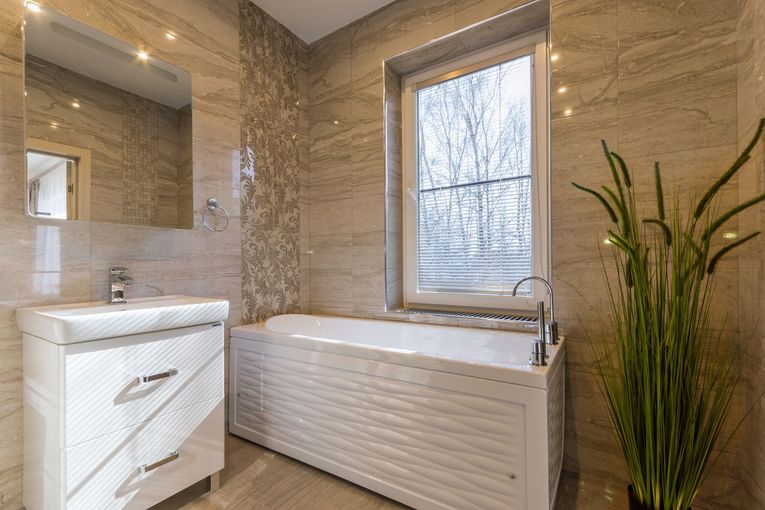 Bright bathroom with white vanity, large tub, beige tile walls, and a tall green plant by the window.